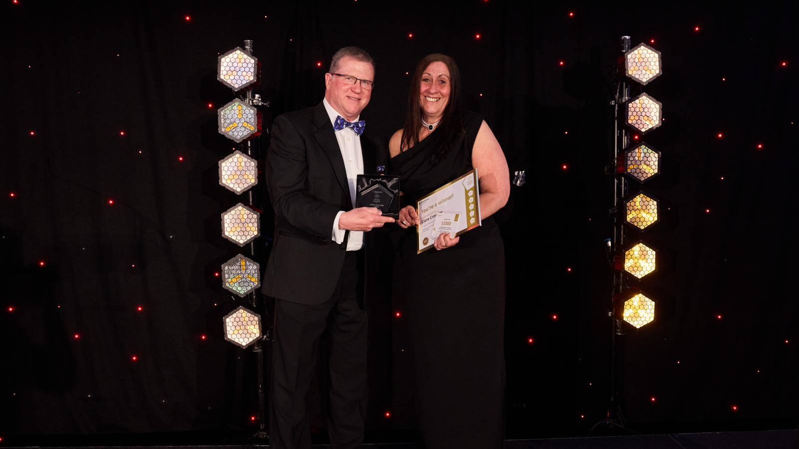 Two people stood on a stage at an award ceremony posing for a photo holding a trophy and certificate