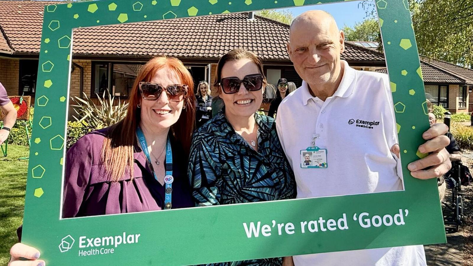 Three people smiling outdoors, holding a green frame with "Exemplar Health Care" and "We're rated 'Good'." Bright, sunny day, joyful atmosphere.