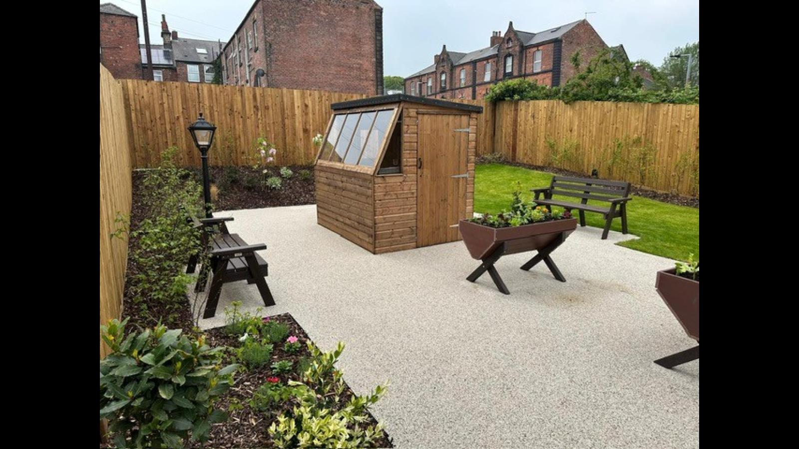 Cozy garden with a wooden shed, seating, and planters. Surrounded by a tall fence, brick buildings in the background. Peaceful and inviting ambiance.