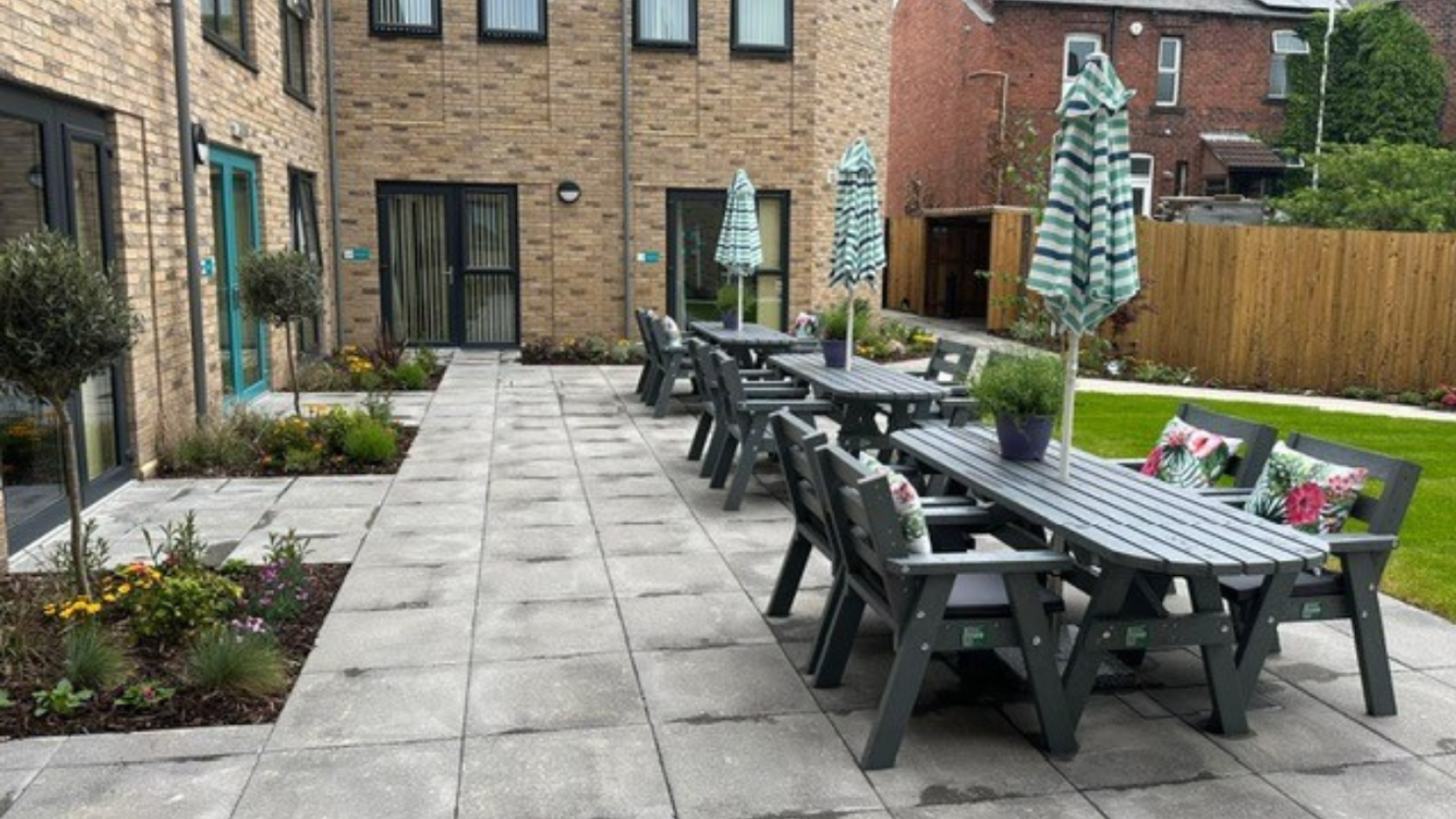 Outdoor patio with gray wooden tables and benches, adorned with floral cushions. Green umbrellas provide shade, surrounded by brick buildings and garden plants.