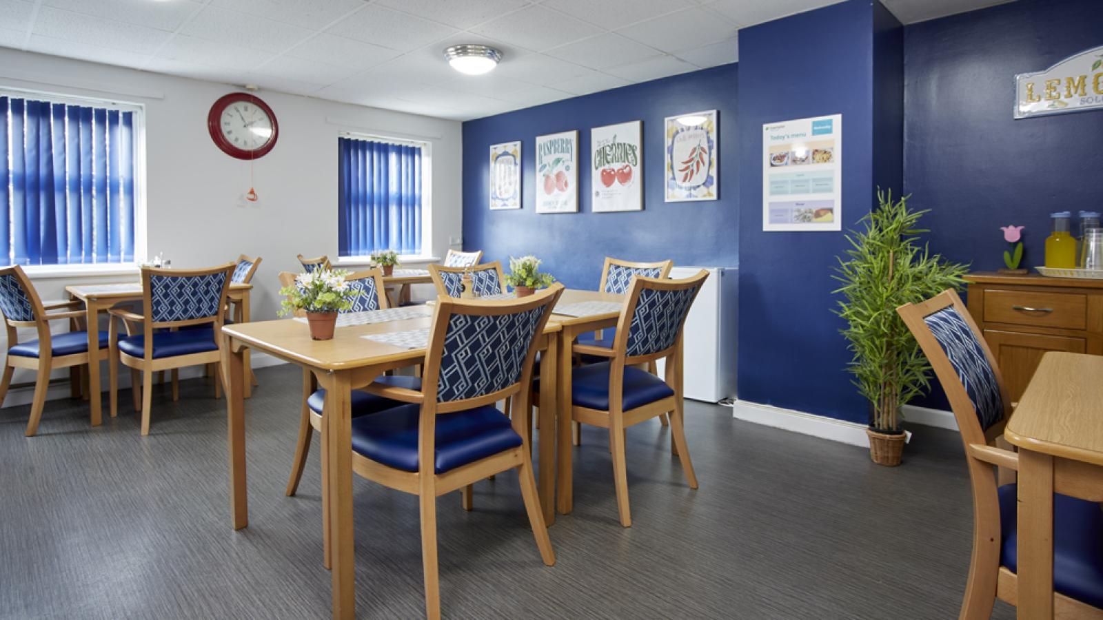 A cozy dining room with blue decor, wooden tables, and chairs. Art and a menu adorn the walls. Plants add a fresh touch