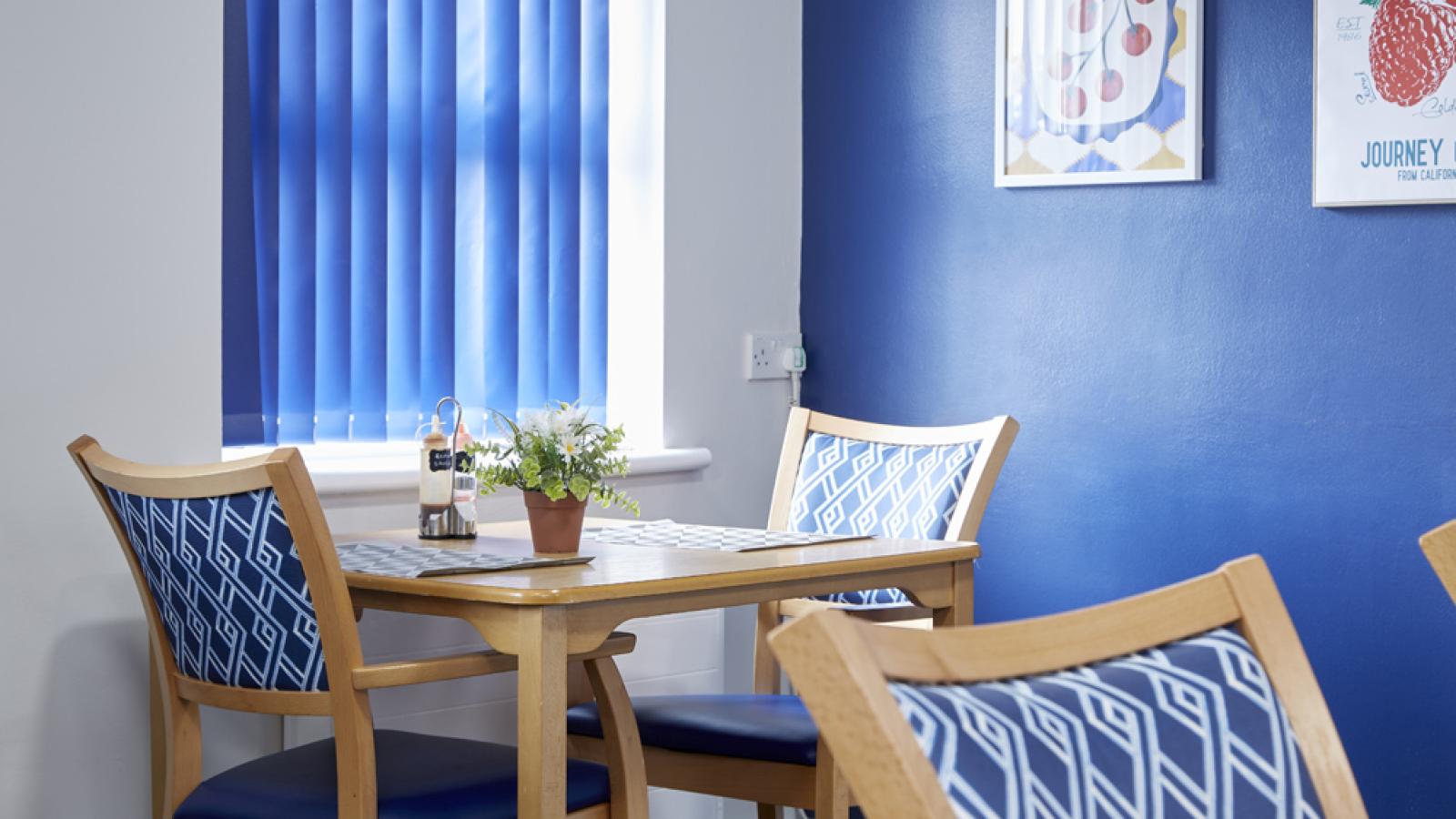 A cozy dining area with blue chairs, a wooden table, and a potted plant. Blue blinds and wall art add a vibrant touch