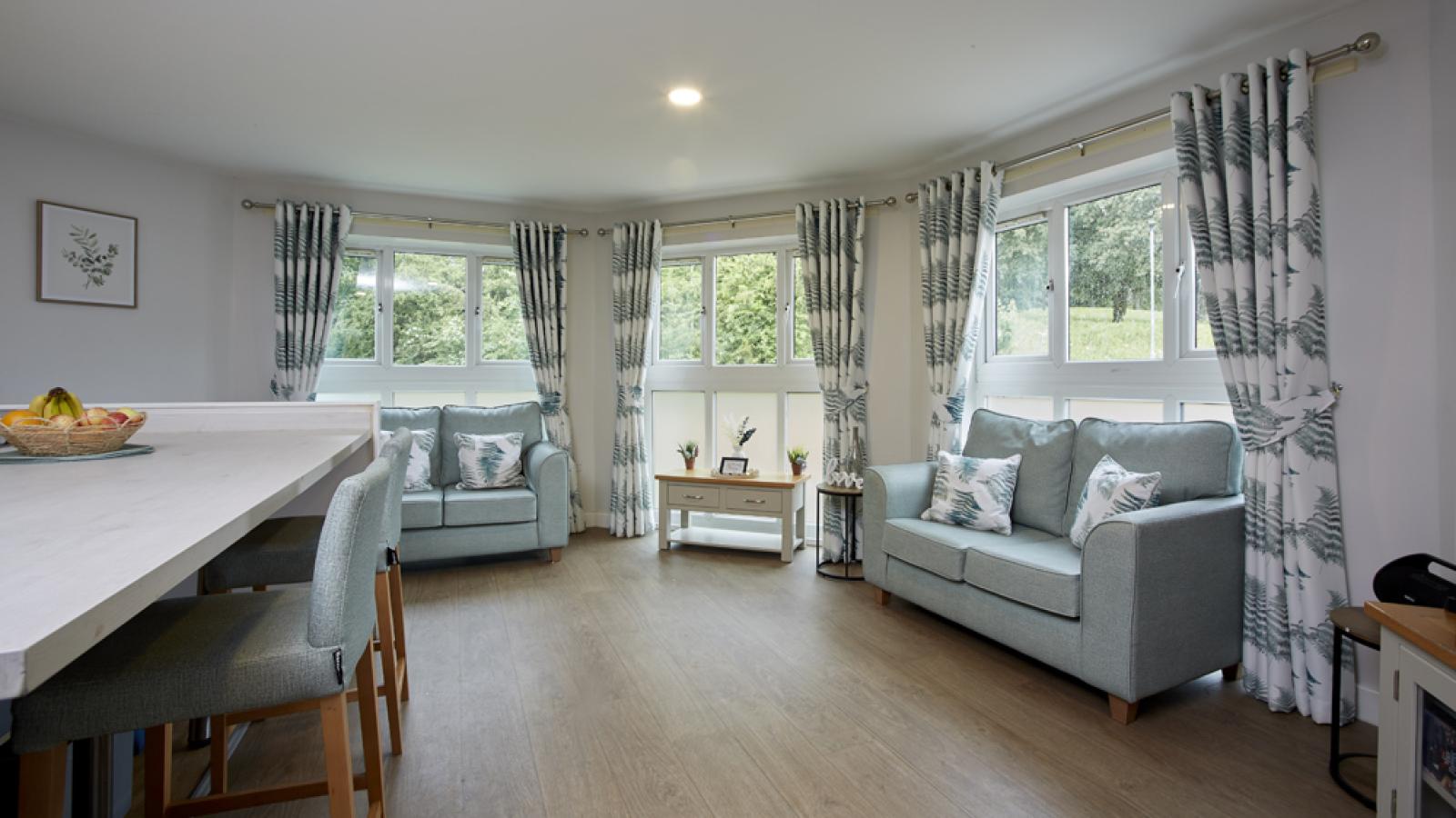 Bright living room with blue sofas, patterned curtains, and large windows overlooking greenery. A fruit bowl sits on the table.
