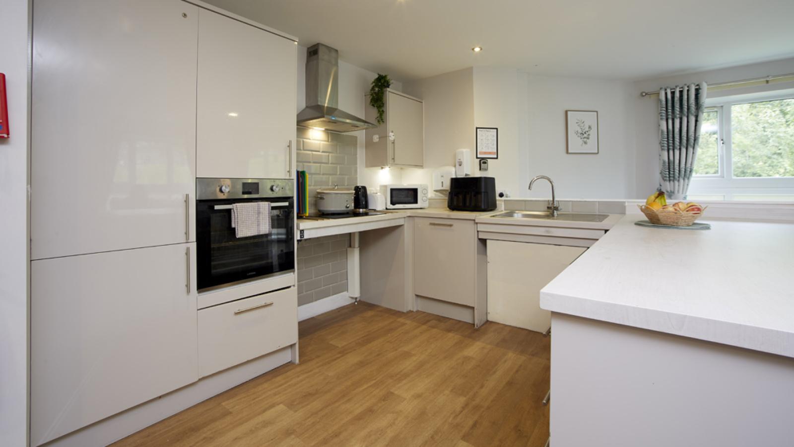 Modern kitchen with white cabinets, wood floor, oven, microwave, and sink. Window view with fruit bowl on counter.