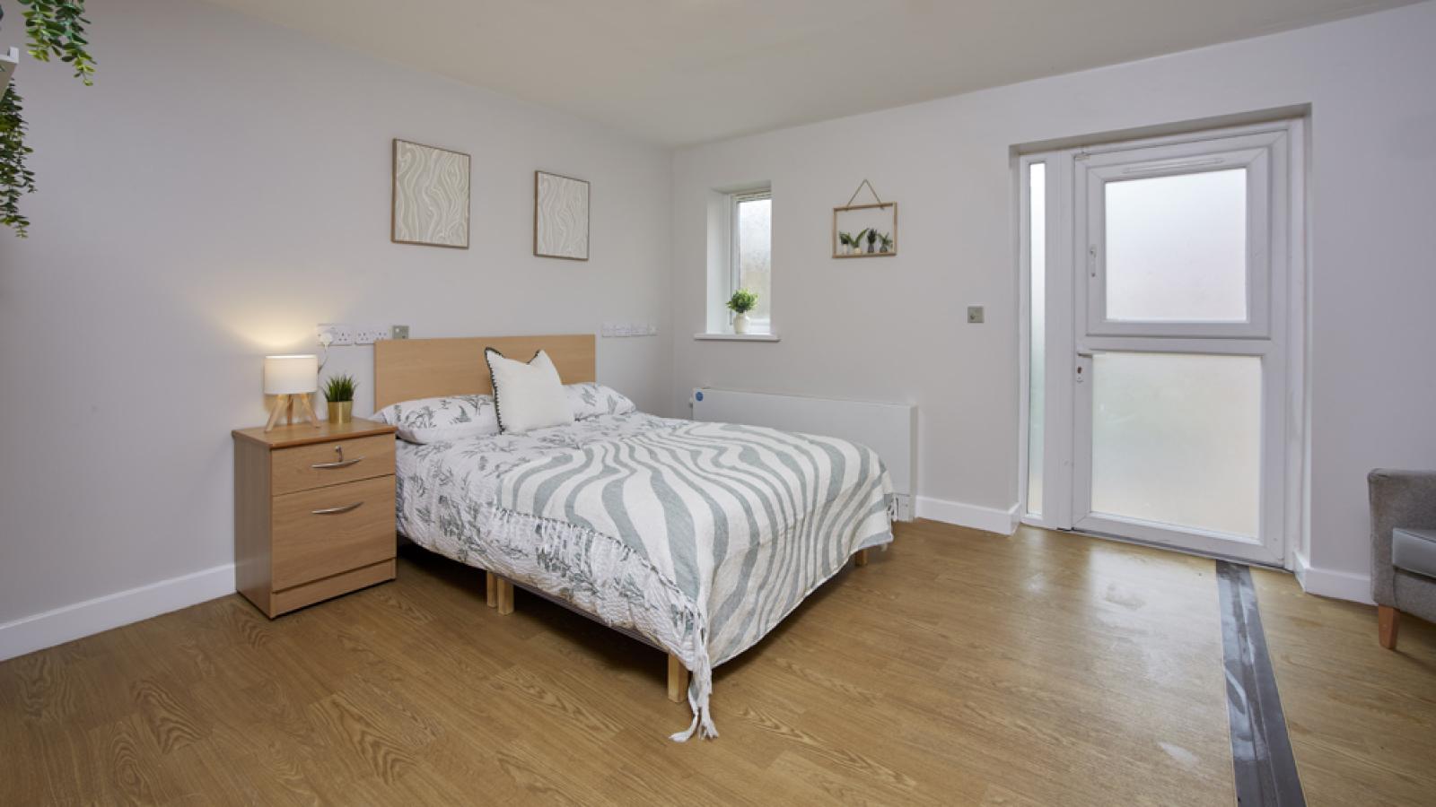 Modern bedroom with a cozy bed, wooden floor, bedside table, and soft lighting. Minimalist decor with plants and framed art.