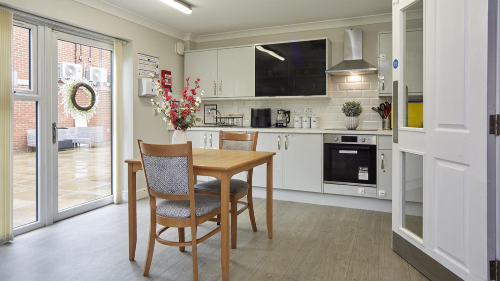 Modern kitchen with white cabinets, wooden table, chairs, and flowers. Glass doors open to a patio with a wreath on the wall