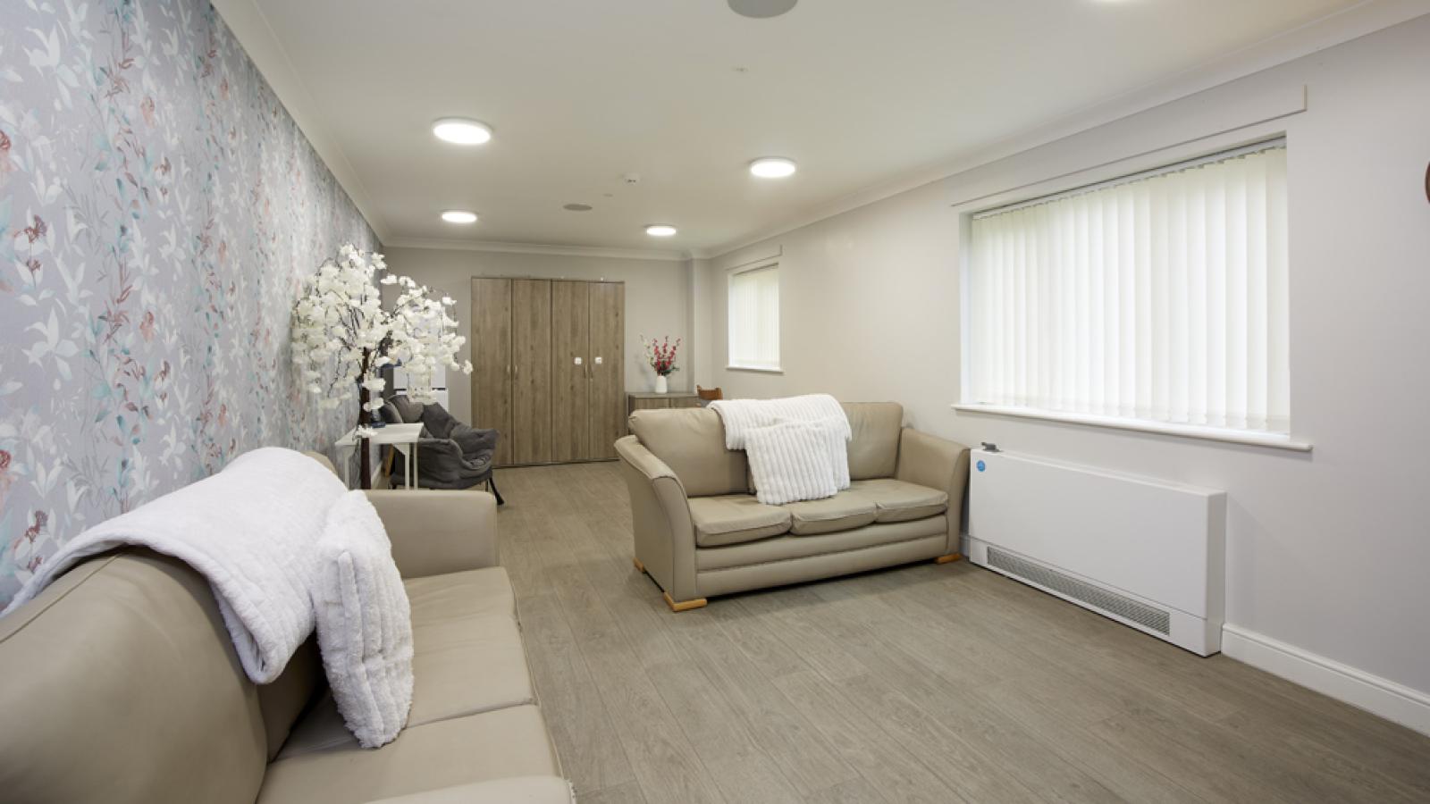 Modern living room with floral wallpaper, wall clock, TV, and cozy sofa. Vertical blinds filter light through the window