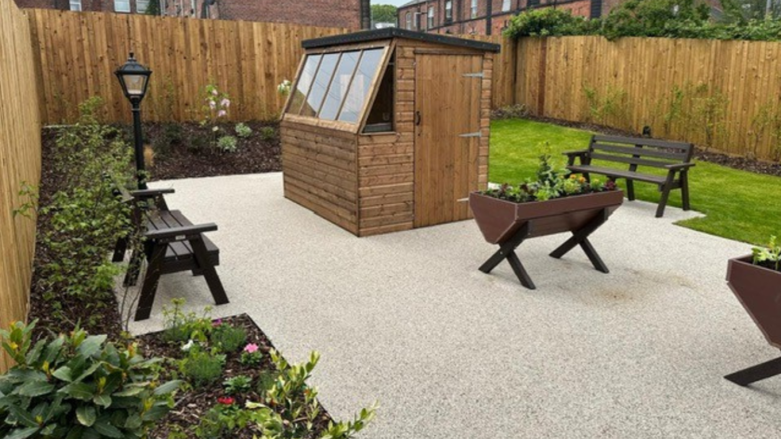 Cozy garden with a wooden shed, seating, and planters. Surrounded by a tall fence, brick buildings in the background. Peaceful and inviting ambiance.