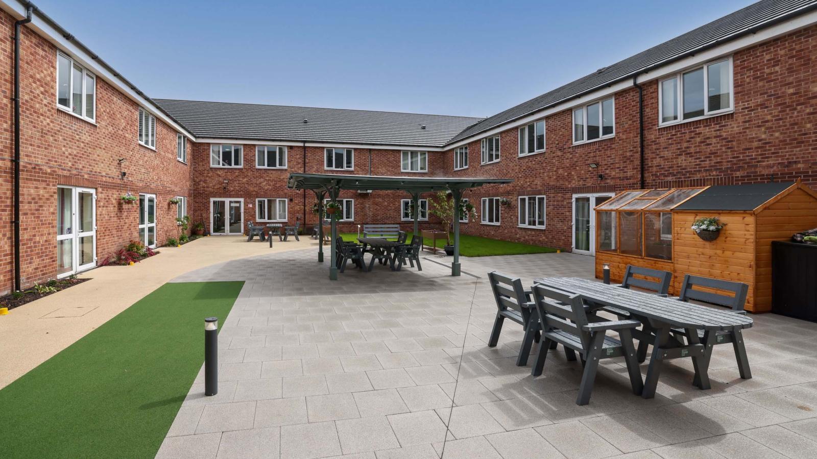 Outdoor courtyard area with patio area and strips of grass, with seating area, benches, and shed with a greenhouse