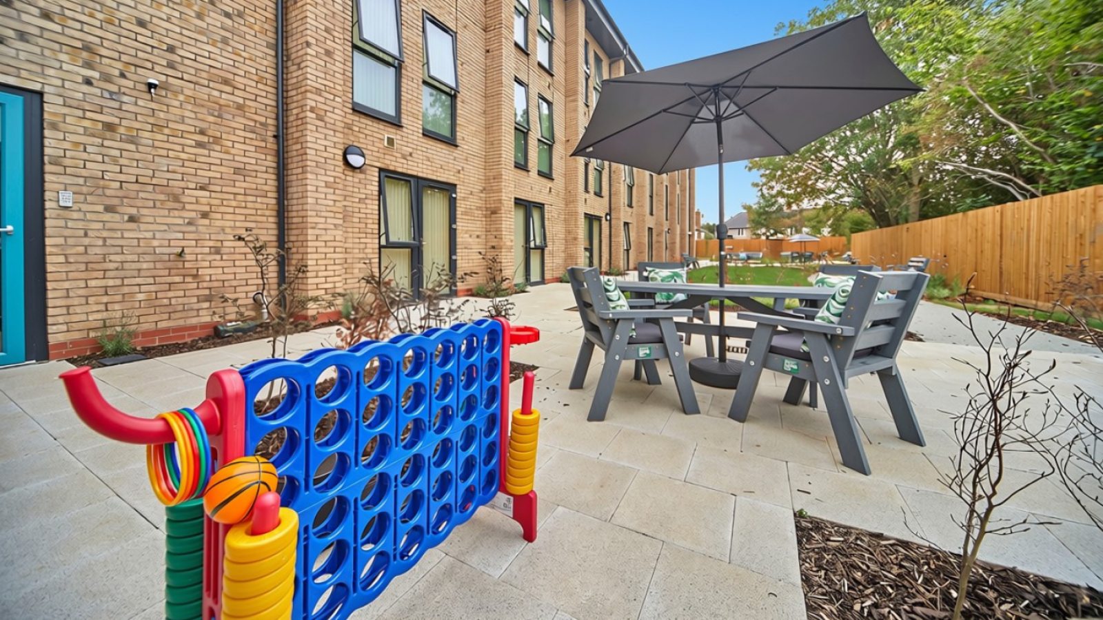 Outdoor courtyard with colorful play panel, picnic table and umbrella, brick building, trees, and fenced patio.