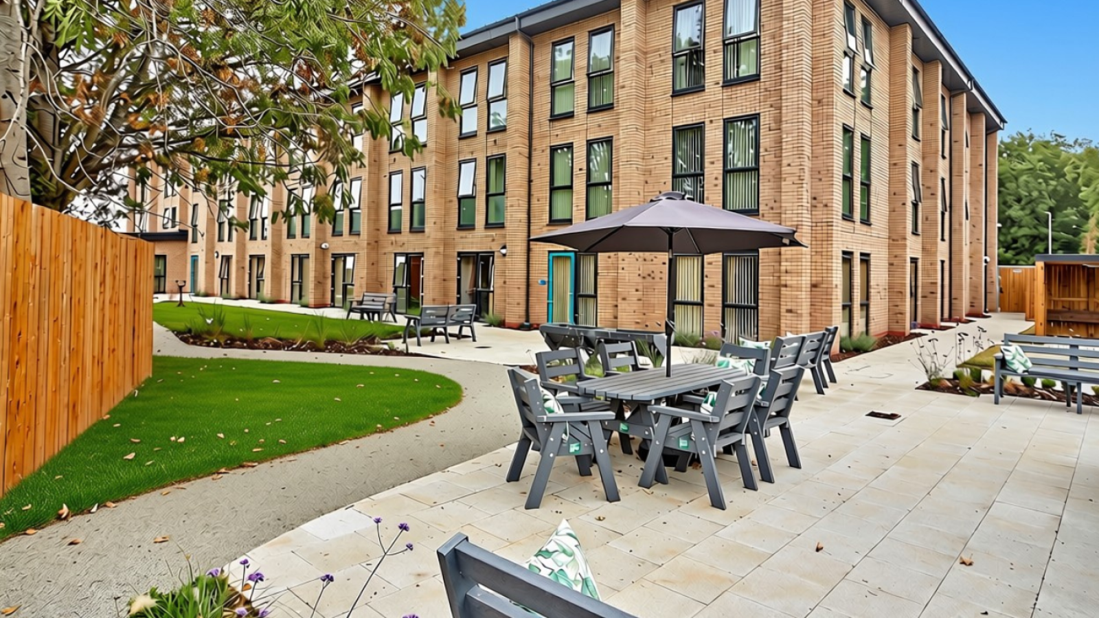 Modern brick building with landscaped courtyard, patio tables, umbrellas, walkways, and lush lawns under a bright blue sky.