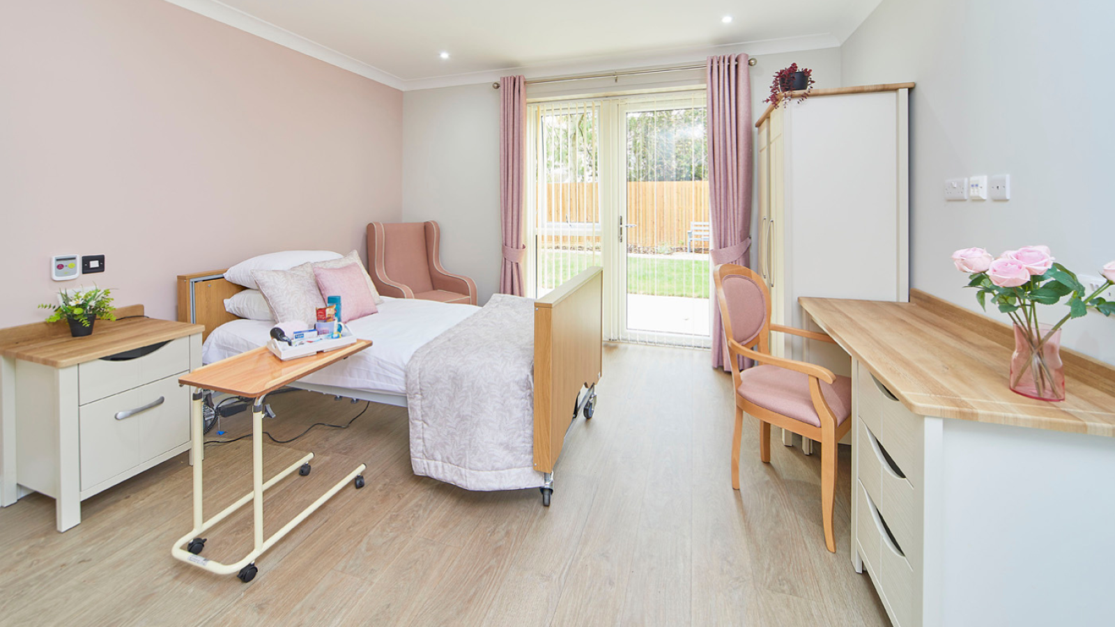 Bright, welcoming care bedroom with an adjustable bed, armchairs, dresser, desk, pink curtains, and garden view through glass doors.