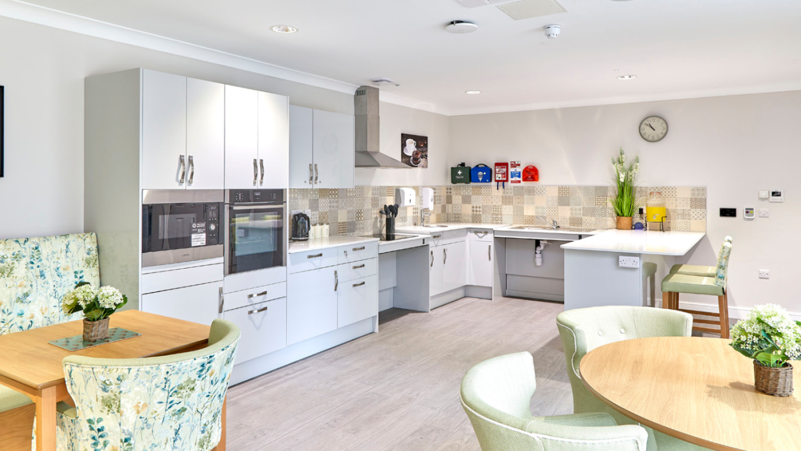 Bright, modern open-plan kitchen and dining area with white cabinets, tiled backsplash, island, wood tables, and pastel chairs; warm, inviting tone.