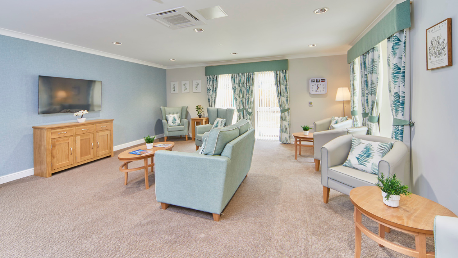 Bright, serene living room with pale blue walls, sofas, armchairs, wood tables, TV console, patterned curtains, and sunlit windows.