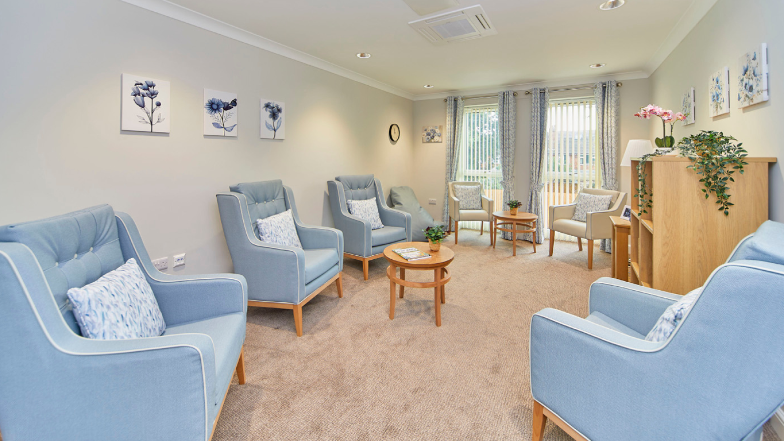 Bright, welcoming lounge with blue armchairs, coffee tables, floral art, plants, and large windows creating a calm, homelike setting.